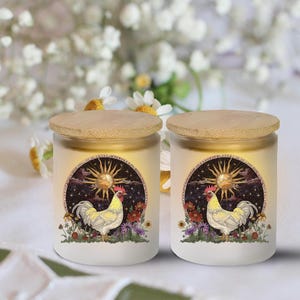 May include: Two white glass candles with bamboo lids. Each candle features a circular design with a sun, rooster, and floral elements. The candles are set against a soft, blurred background of white flowers.