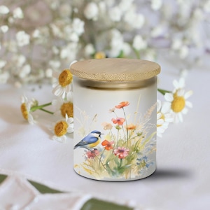 May include: A cylindrical jar with a bamboo lid. The jar features a watercolor painting of a blue bird perched among wildflowers in shades of orange, pink, and green. The background is blurred with white flowers.