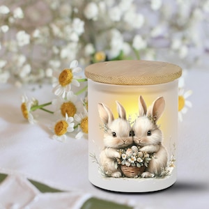 May include: A white candle holder with a wooden lid features a watercolor illustration of two bunnies holding a basket of eggs and flowers. The candle holder is surrounded by white flowers.
