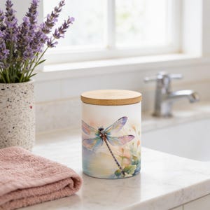 May include: A cylindrical white ceramic canister featuring a colorful watercolor-style dragonfly illustration on the front. The container is topped with a circular light-colored wooden lid, providing a natural contrast to the smooth white surface.