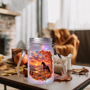 May include: A frosted glass mason jar lantern featuring a printed sunset landscape with a horse, autumn trees, and mountains. The jar has a silver metal lid and a thick, natural-colored rope handle, illuminated by small, colorful LED lights.