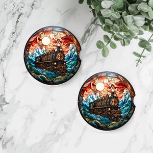 May include: Two round coasters with a stained glass design featuring a train traveling through a mountain range with a sunset in the background. The coasters are white with a glossy finish.