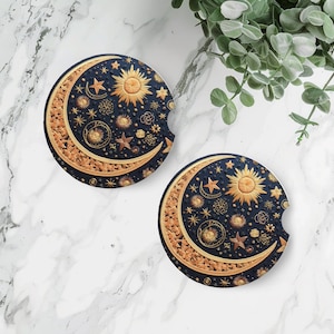 May include: Two round, black coasters with a gold crescent moon and sun design. The design features a crescent moon with stars and a sun with rays. The coasters are on a white marble surface.