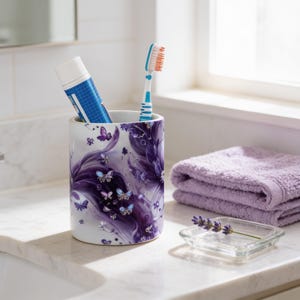 May include: A cylindrical ceramic toothbrush holder featuring a white base with a purple swirling pattern and small butterfly illustrations. The holder contains a blue and white toothpaste tube and a white toothbrush with blue and orange bristles.