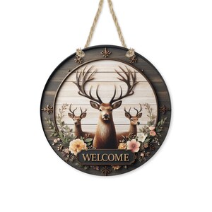 Op de afbeelding: Ronde houten welkomstbord met drie herten met grote geweien, omringd door bloemen. Het bord heeft een donkerbruine rand en een lichtbruine achtergrond met het woord "WELCOME". Er is een touw bevestigd om op te hangen.