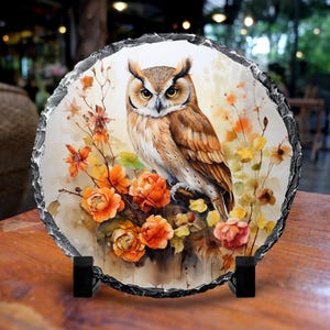 May include: A round, decorative stone featuring a watercolor painting of a brown owl perched on a branch with orange and yellow flowers. The stone has a rough, textured edge and is displayed on a black stand. The artwork has a natural, rustic theme.