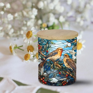 May include: A cylindrical container with a bamboo lid. The container's body features a stained-glass design of two birds perched on a branch with red berries, set against a blue sky and foliage. The background includes white flowers.