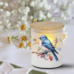 May include: A white candle holder with a wooden lid, featuring a bluebird illustration perched on a branch with berries. The candle holder is cylindrical and has a soft, warm glow.