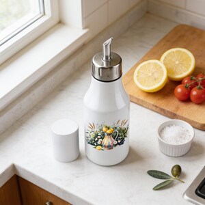 May include: A white oil dispenser bottle with a stainless steel pour spout and a matching white cap. The bottle features a colorful graphic illustration of a pitcher surrounded by lemons, wheat stalks, and small white flowers on the front surface.