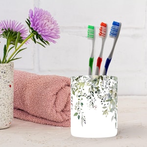 May include: A white toothbrush holder with a watercolor floral design, holding three toothbrushes with green, red, and blue bristles. A pink towel and a vase with purple flowers are also in the image.