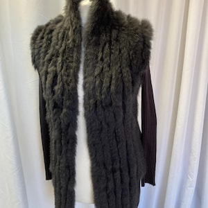 May include: A dark gray fur vest with long sleeves. The vest has a fluffy texture and a long, open front. The sleeves are a dark burgundy color. The garment is displayed on a mannequin against a white backdrop.