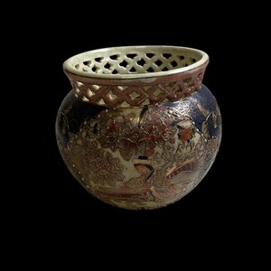 Op de afbeelding: Een decoratieve keramische vaas met een ronde body en een patroonrand. De vaas heeft een gedetailleerd ontwerp met bloemen- en landschapselementen in bruin, goud en blauw. De rand heeft een rasterpatroon.