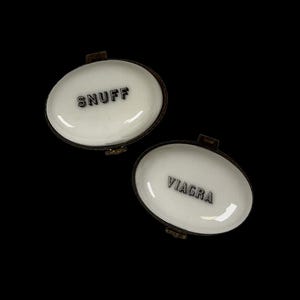 May include: Two oval-shaped metal boxes with hinged lids. The top box features a white enamel surface with the black text SNUFF. The bottom box is open, revealing a white enamel interior with the black text VIAGRA embossed on the base surface.