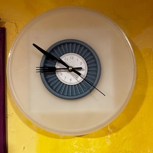 May include: A round wall clock with a white face and a blue center. The clock hands are black, with the minute hand shaped like a fork. The clock is mounted on a yellow wall.