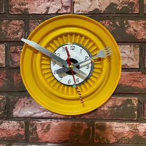 May include: A unique yellow clock with a white clock face and silver hands. The clock hands are a fork and knife. The clock is mounted on a brick wall. The clock face is surrounded by a yellow plastic ring.