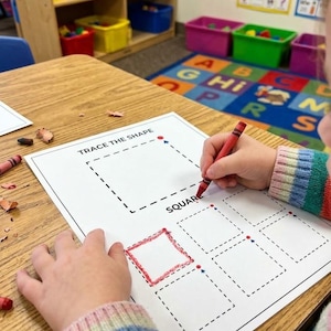 May include: A white worksheet titled TRACE THE SHAPE with dashed square outlines and the word SQUARE printed on it. A red crayon is being used to trace one of the squares on the paper, which is resting on a wooden table surface.