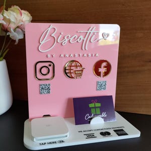 May include: A pink acrylic sign with the text "Biscotti by Anastasia" in white lettering. The sign has icons for Instagram, a globe with a shopping cart, and Facebook. The sign also has a white square with a QR code. Below the sign is a white base with the text "Tap Here" and a white square with a black QR code. The base also has the text "We accept contactless payments" and a Visa logo.