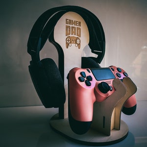 May include: A wooden stand with the words "Gamer Dad" engraved on it. The stand holds a pair of black headphones and a pink video game controller.