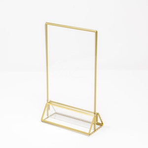 May include: A clear acrylic sign holder with a gold trim and a triangular base. The holder is designed to display a sign or menu.