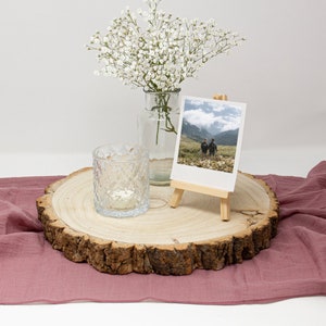 May include: A wooden slice with a rustic bark edge, a clear glass candle holder, a small easel with a photo of two people standing in a field, and a vase with white baby's breath flowers on a pink fabric surface.