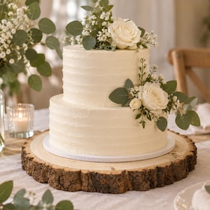 May include: A two-tier white frosted cake with horizontal textured lines, topped and decorated with white roses, baby's breath, and eucalyptus leaves, resting on a round natural wood slice with visible bark edges on a table.