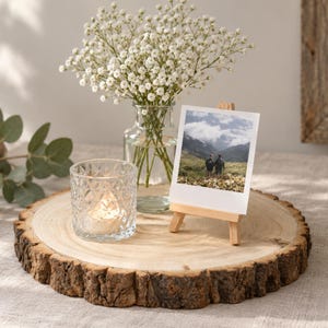 May include: A round wooden slice with natural bark edges holding a clear glass vase filled with white baby's breath flowers, a small clear textured glass candle holder with a lit tea light, and a square photograph displayed on a miniature wooden easel.