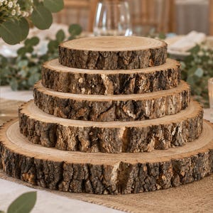 May include: A tiered display stand composed of five circular wood slices stacked in descending order of size. Each slice features a smooth, light-colored top surface with visible grain rings and a rough, dark brown natural bark edge.