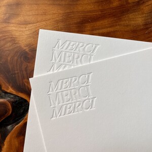 K&ouml;nnte beinhalten: Zwei wei&szlig;e Karten mit dem Wort "MERCI" in einer fetten, serifenlosen Schriftart gepr&auml;gt. Das Wort wird dreimal auf jeder Karte wiederholt.