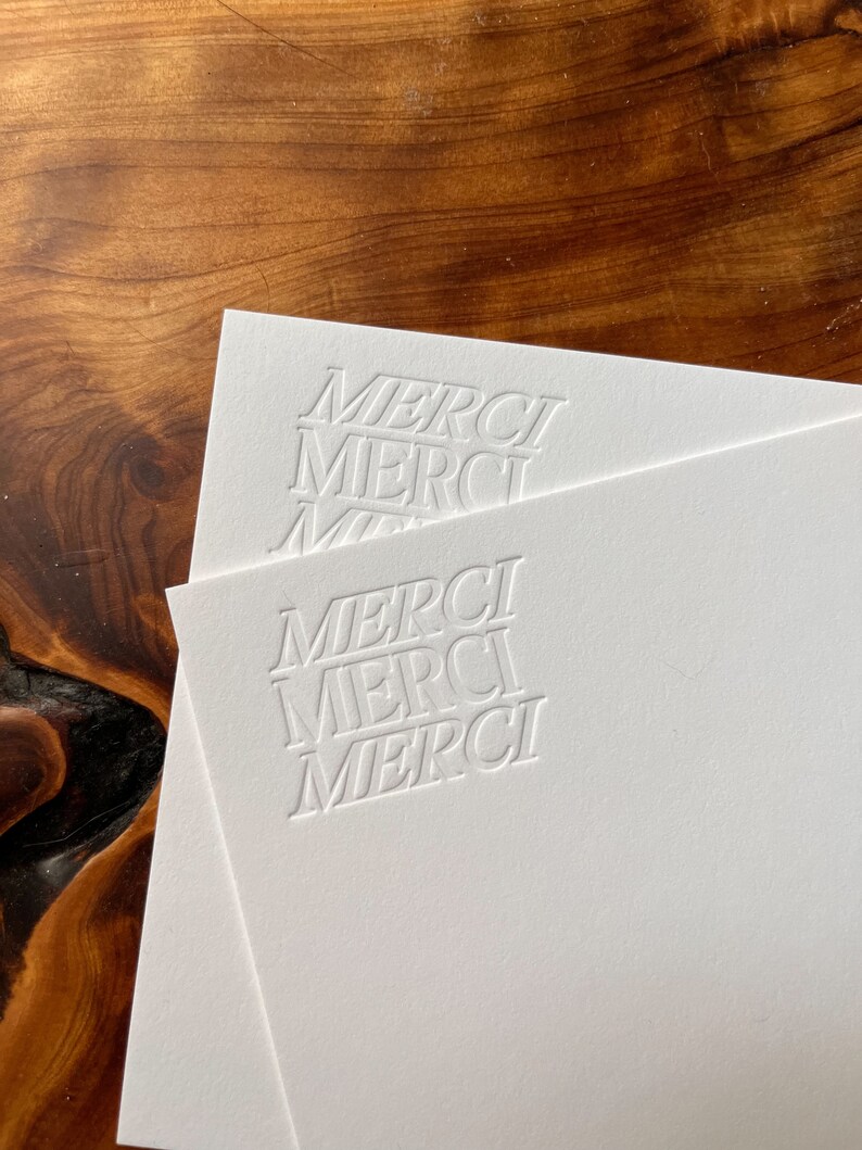K&ouml;nnte beinhalten: Zwei wei&szlig;e Karten mit dem Wort "MERCI" in einer fetten, serifenlosen Schriftart gepr&auml;gt. Das Wort wird dreimal auf jeder Karte wiederholt.