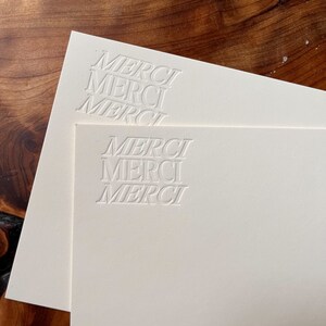K&ouml;nnte beinhalten: Zwei wei&szlig;e Karten mit dem Wort "MERCI" in einer stilisierten Schriftart gepr&auml;gt. Die Karten liegen &uuml;bereinander.