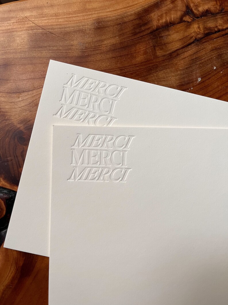 K&ouml;nnte beinhalten: Zwei wei&szlig;e Karten mit dem Wort "MERCI" in einer stilisierten Schriftart gepr&auml;gt. Die Karten liegen &uuml;bereinander.