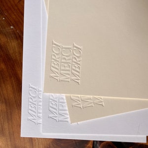 K&ouml;nnte beinhalten: Drei wei&szlig;e Karten aus Karton mit dem Wort "MERCI" in einem gestapelten, vertikalen Design gepr&auml;gt. Die oberste Karte ist cremefarben.