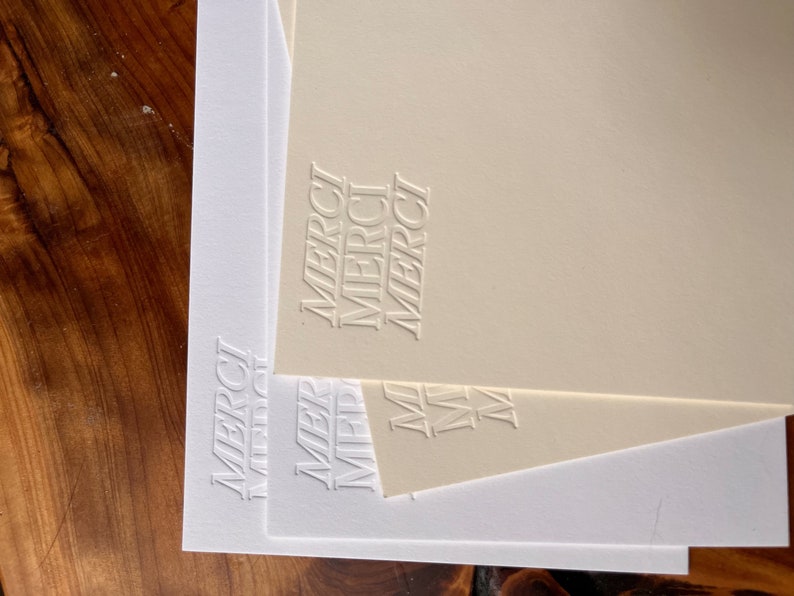 K&ouml;nnte beinhalten: Drei wei&szlig;e Karten aus Karton mit dem Wort "MERCI" in einem gestapelten, vertikalen Design gepr&auml;gt. Die oberste Karte ist cremefarben.