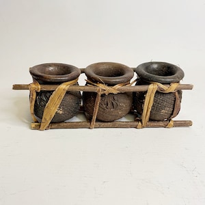 May include: Three small brown clay pots with a textured surface are arranged in a row on a wooden frame with woven straw accents.