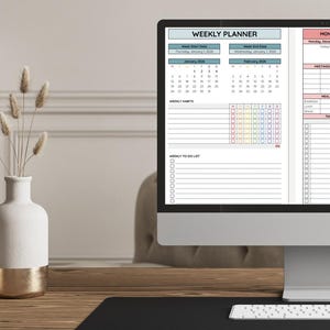 May include: A computer monitor displaying a digital weekly planner with sections for meetings, meal plans, and tasks, alongside two white and gold ceramic vases containing dried grass stems on a wooden desk with a white keyboard.