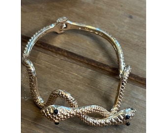 May include: Gold-toned snake bracelet with textured scales and black gem eyes. The bracelet features a hinged clasp and a coiled snake design, creating a unique and stylish accessory. The bracelet is shown on a wooden surface.