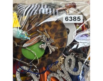 May include: A close-up of a variety of jewelry and accessories in a clear plastic bag. The bag contains a mix of colorful beads, charms, and pendants, including a silver cross pendant, a green and white striped bead, and a brown and black leopard print charm. The bag also includes a tag with the number "6385" on it.