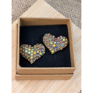 May include: Two heart-shaped earrings with iridescent rhinestones are displayed in a small, open, brown cardboard box. The earrings have a gold-colored metal base and are set against a black background.