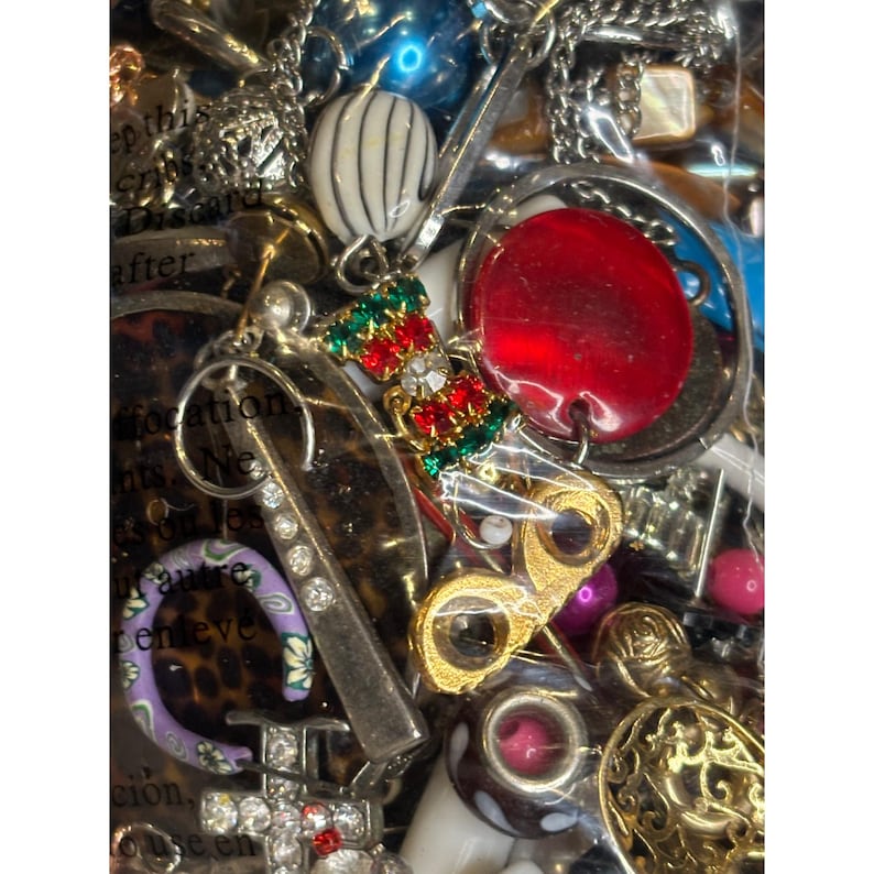 May include: A close-up of a variety of colorful beads, charms, and jewelry pieces in a clear plastic bag. The items include a red round bead, a gold charm with a pair of glasses, a silver charm with rhinestones, and a green and red charm with a Christmas stocking design.