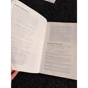 Op de afbeelding: Open boek met tekst op de pagina's. De linkerpagina heeft de titel "Chapter Organization" en bevat hoofdstuktitels. De rechterpagina bevat bronnen voor instructeurs. Het boek ligt open op een donkere ondergrond.