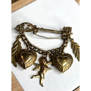 May include: Vintage gold-tone brooch featuring a safety pin design with a chain holding two heart-shaped charms and a small cherub. The brooch also includes two leaf charms. The brooch is a romantic accessory.