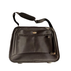 Puede incluir: Maletín rodante negro con bandolera. El maletín tiene un bolsillo frontal con cremallera y un pequeño logotipo dorado. La bolsa tiene una bandolera acolchada y dos ruedas en la parte inferior.