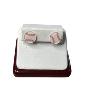 May include: A pair of baseball-themed earrings on a white display stand. The earrings are made of clear resin with a red and white baseball design.