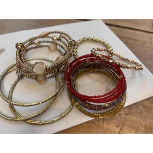May include: A set of six gold and red beaded bracelets. The bracelets are stacked on top of each other and feature different patterns and textures. Some of the bracelets have small, round, white stones.