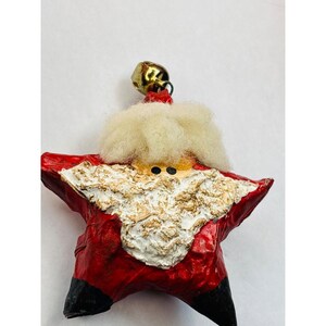 May include: A star-shaped Christmas ornament depicting Santa Claus. The ornament features a red body with black tips, a white beard, and a gold bell at the top. The beard and mustache are textured, and the face has two small black eyes.