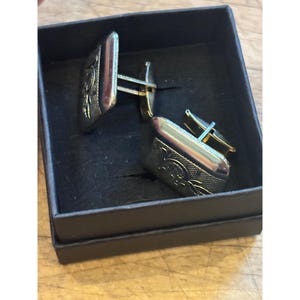 Puede incluir: Un par de gemelos plateados con diseño rectangular y detalles florales, presentados en una caja negra. Los gemelos tienen una superficie texturizada y un acabado pulido, ideales para ocasiones formales.