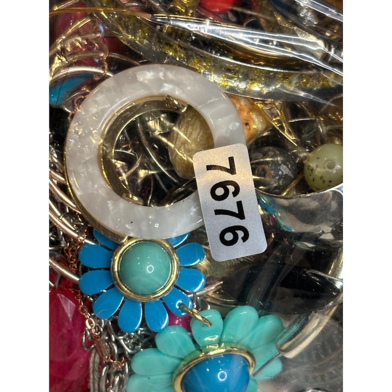 May include: A close-up of a variety of jewelry pieces, including a white and gold ring, blue and turquoise flower-shaped earrings, and a variety of beads and charms. The jewelry is displayed in a clear plastic bag with the number 7676 printed on it.