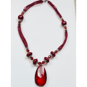 May include: A red beaded necklace with a large, teardrop-shaped pendant. The necklace features multiple strands of small red beads, interspersed with larger, round beads and silver-toned accents. The pendant is a faceted, deep red gemstone.