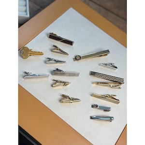 Puede incluir: Una colección de 11 clips de corbata de oro y plata. Los clips de corbata están dispuestos en dos filas sobre una superficie marrón claro. Algunos de los clips de corbata tienen diseños decorativos, mientras que otros son lisos.