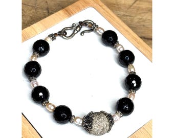 May include: A beaded bracelet featuring faceted black beads, small pearls, and a central silver-toned bead with a textured surface. The bracelet is secured with a silver-toned clasp.